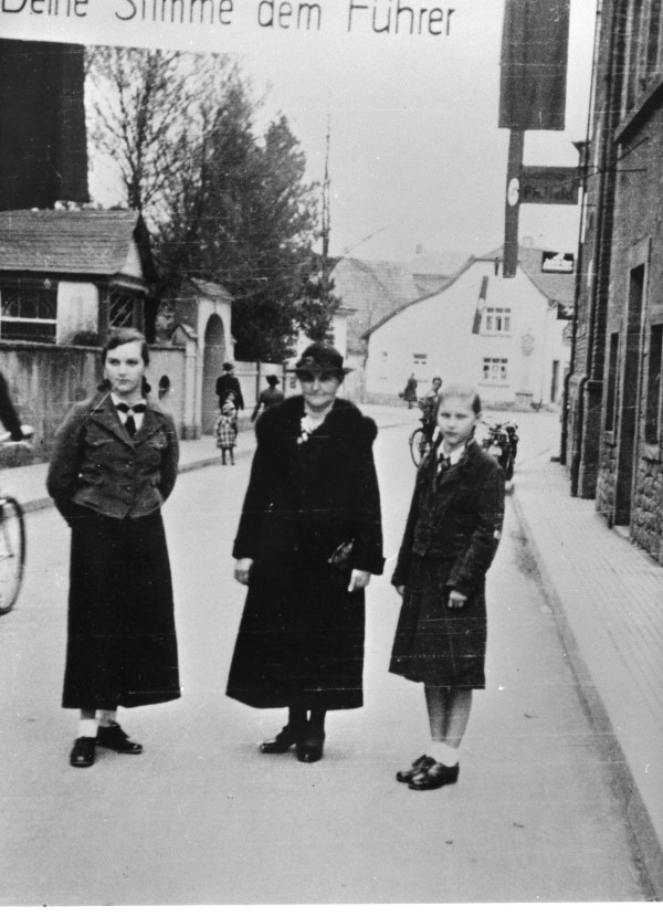 Wahlkampf in Lohr 1938 – Die Vorstadtstrae mit dem Spruchband „Deine Stimme dem Fhrer“, darunter eine Mutter mit ihren Tchtern, diese in der Uniform der „Jungmdel“ bzw. des „BDM“.