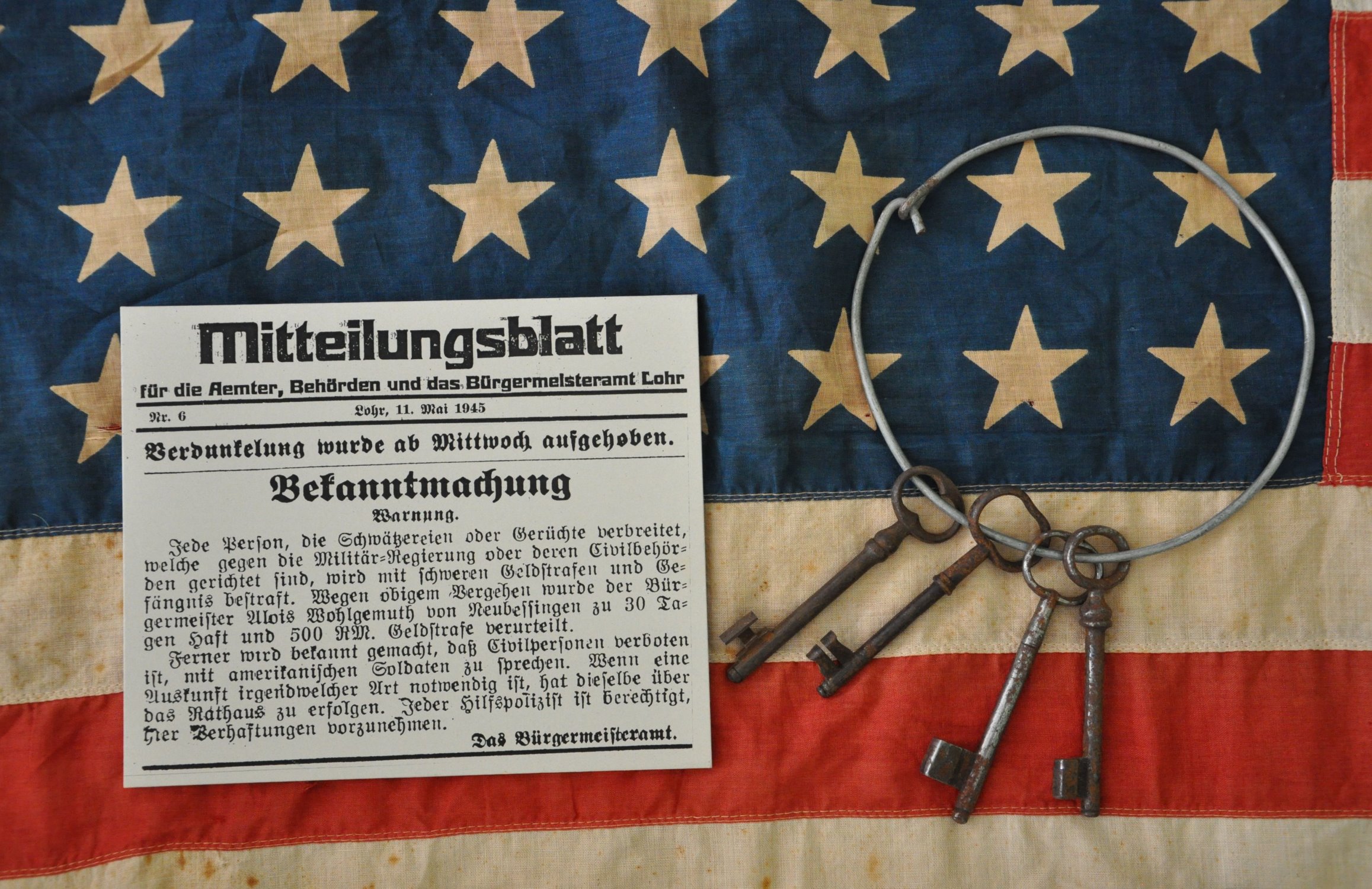 US-Sternenbanner, Bekanntmachung („Warnung“) im „Mitteilungsblatt fr die Aemter, Behrden und das Brgermeisteramt Lohr“, Nr. 6 vom 11. Mai 1945 und Schlsselbund als Hinweis auf drohende Gefngnisstrafen bei Vergehen gegen die Anordnungen der US-Militrregierung (Darstellung in einer Ausstellungsvitrine im Schulmuseum)
